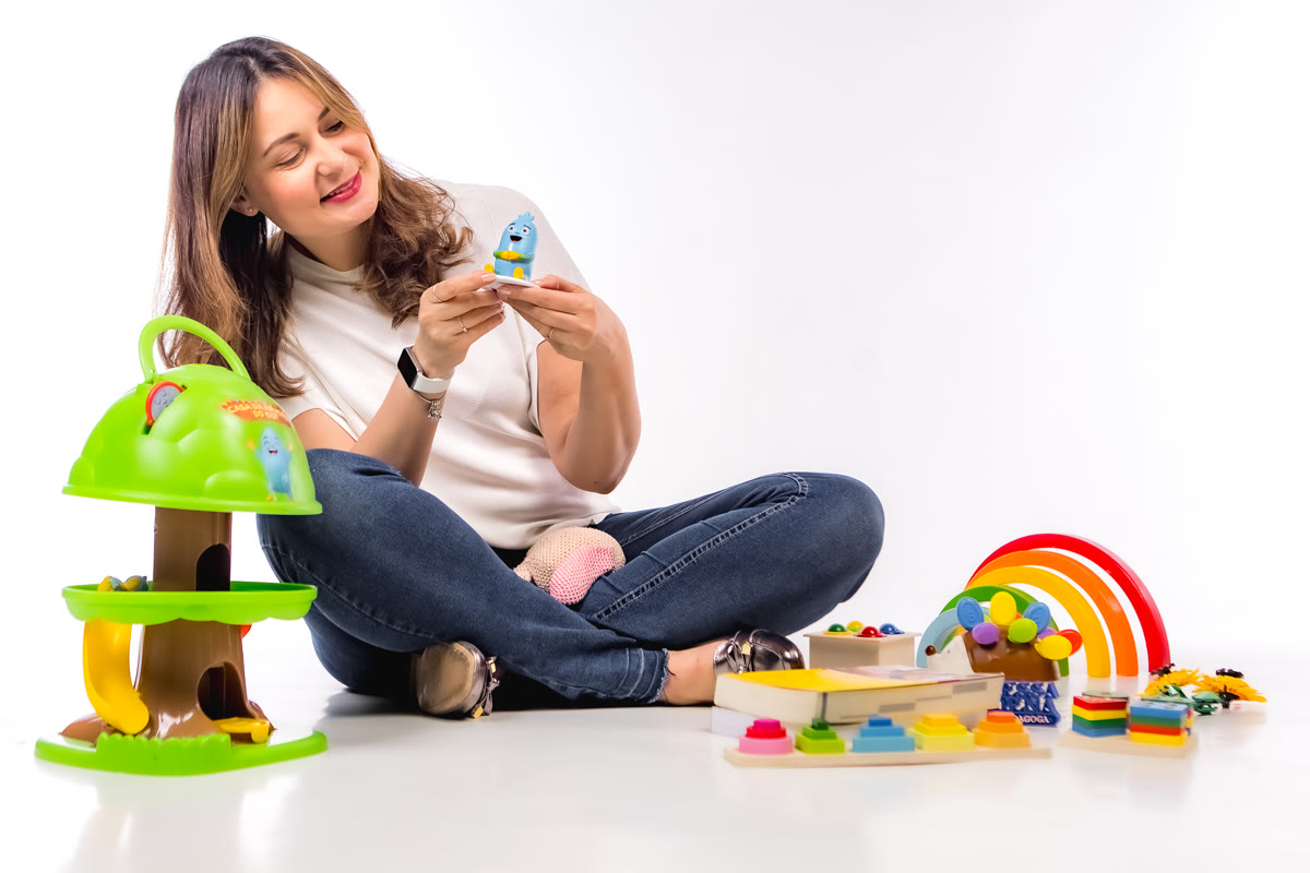 Vanessa sentada no chão olhando o boneco do Gui, com a casa da árvore do Gui, um arco-íris de madeira e outros brinquedos pedagógicos